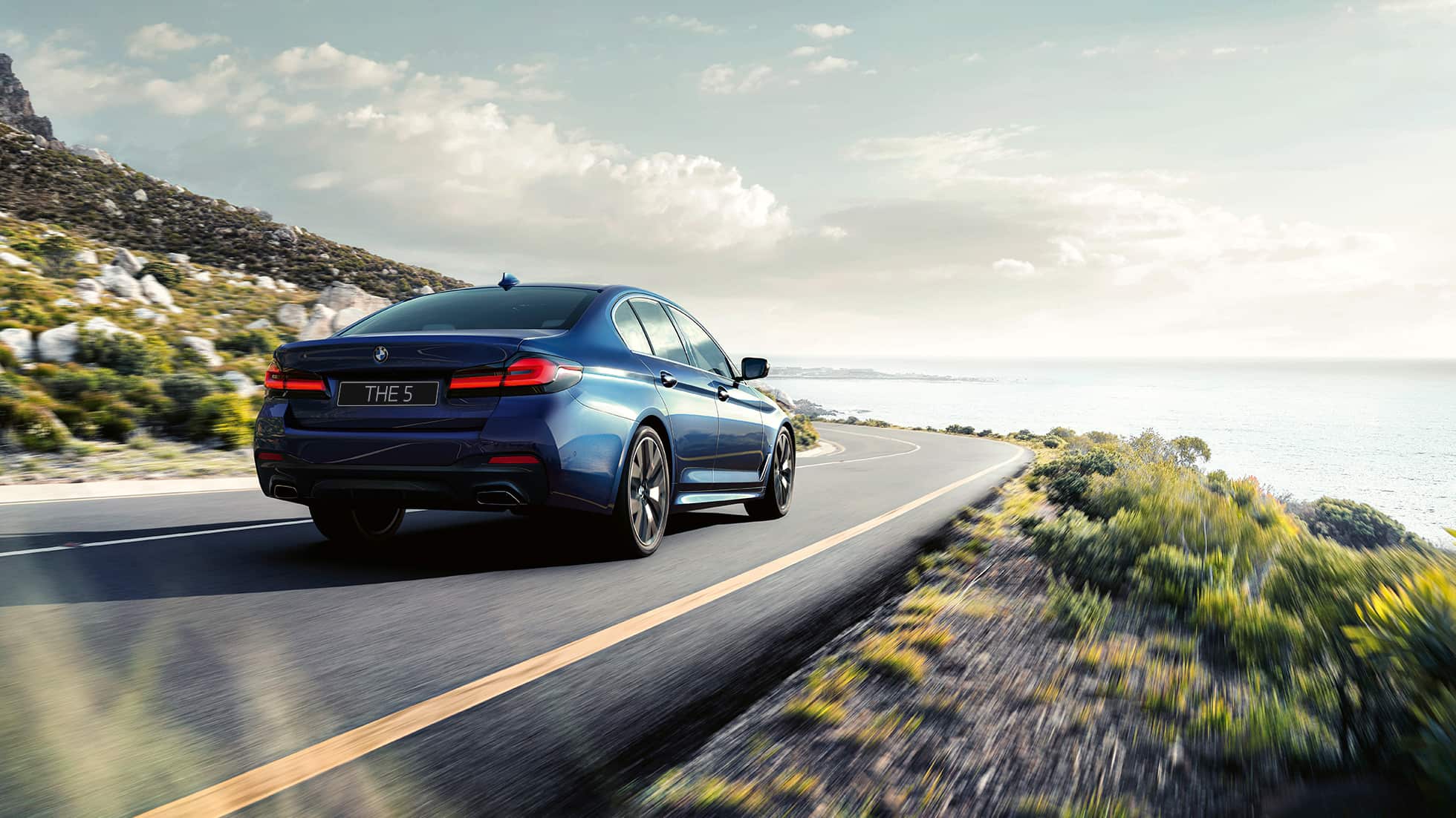 Динамичный BMW 5 серии G30 Динамичный BMW 5 серии G30, цвет «Синий Фитоник», вид сзади с поворотом в три четверти, в движении