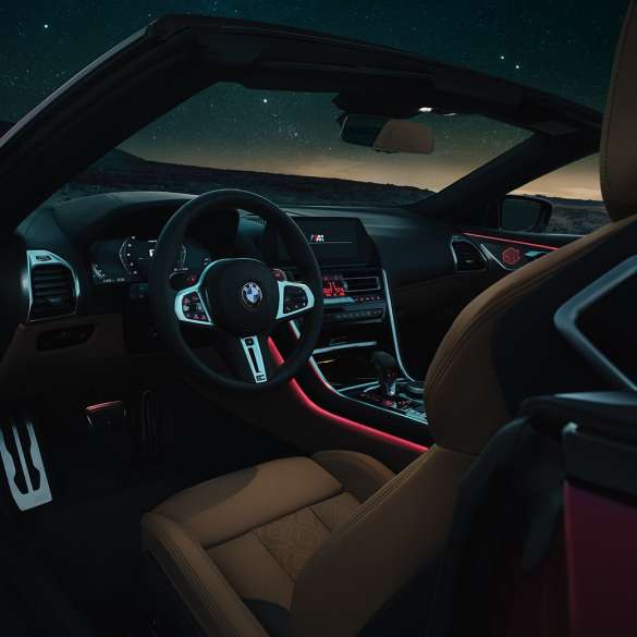 BMW M8 Competition Cabrio, интерер. BMW M8 Competition Cabrio, кокпит с обтянутым кожей рулевым колесом M и фоновой подсветкой красного цвета в темное время суток.