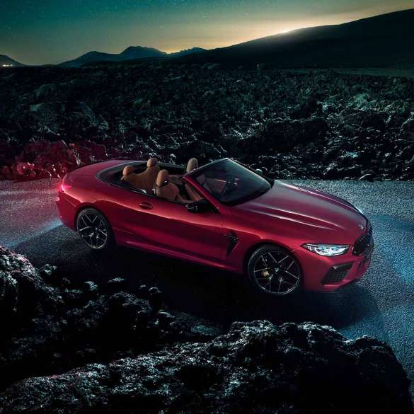 BMW M8 Competition Cabrio, вид сбоку с поворотом в три четверти. BMW M8 Competition Cabrio с открытым мягким верхом, Красный Мотеги металлик, вид сбоку с поворотом в три четверти, неподвижный автомобиль на асфальте в темное время суток.