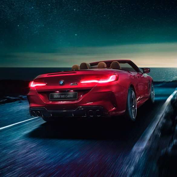 BMW M8 Competition Cabrio, вид сзади с поворотом в три четверти. BMW M8 Competition Cabrio с открытым мягким верхом, Красный Мотеги металлик, вид сзади с поворотом в три четверти, автомобиль движется вдоль побережья в темное время суток.