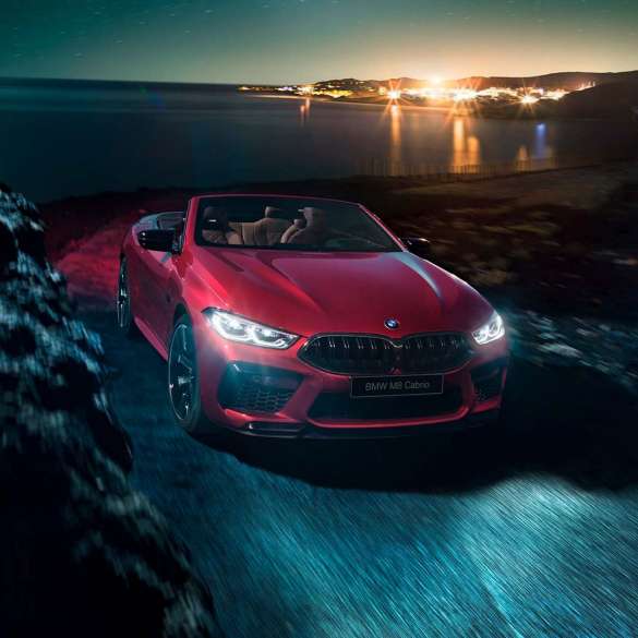 BMW M8 Competition Cabrio, вид спереди с поворотом в три четверти. BMW M8 Competition Cabrio с открытым мягким верхом, Красный Мотеги металлик, вид спереди с поворотом в три четверти, автомобиль движется вдоль побережья в темное время суток.