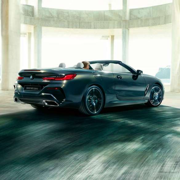BMW M850i xDrive Cabrio, вид сзади с поворотом в три четверти. BMW M850i xDrive Cabrio с открытым мягким верхом, Серый Дравит металлик, вид сзади с поворотом в три четверти, автомобиль движется внутри здания из бетона.