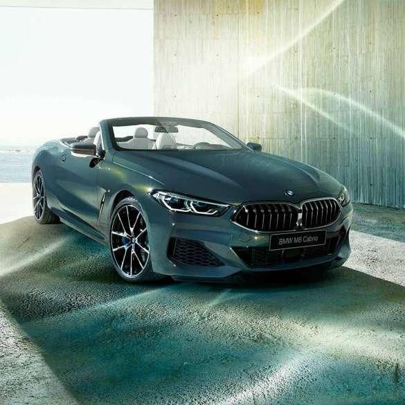 BMW M850i xDrive Cabrio, вид спереди с поворотом в три четверти. BMW M850i xDrive Cabrio с открытым мягким верхом, Серый Дравит металлик, вид спереди с поворотом в три четверти, неподвижный автомобиль на фоне побережья.