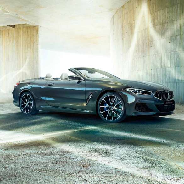 BMW M850i xDrive Cabrio, вид сбоку с поворотом в три четверти. BMW M850i xDrive Cabrio с открытым мягким верхом, Серый Дравит металлик, вид сбоку с поворотом в три четверти, неподвижный автомобиль перед зданием из бетона.