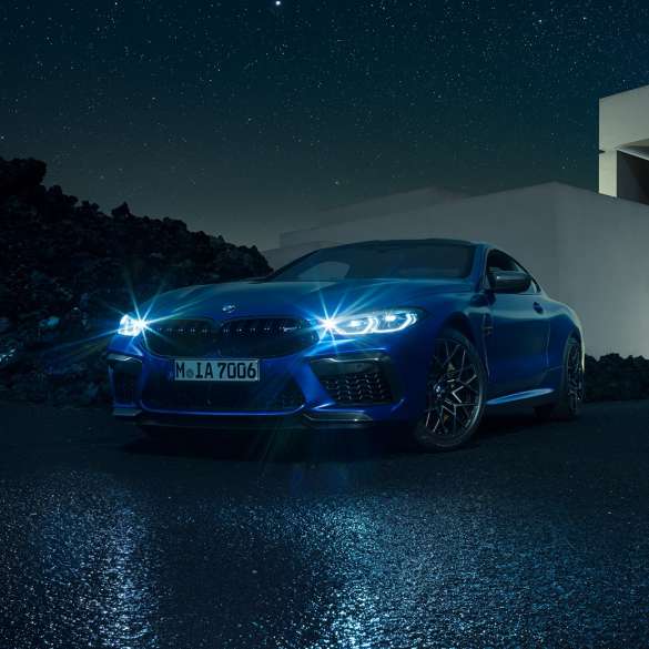 BMW M8Coupe Competition, вид спереди с поворотом в три четверти. BMW M8 Coupe Competitionс включенными светодиодными фарами, Синий Марина Бэй металлик, вид спереди с поворотом в три четверти, неподвижный автомобиль перед виллой в темное время суток.