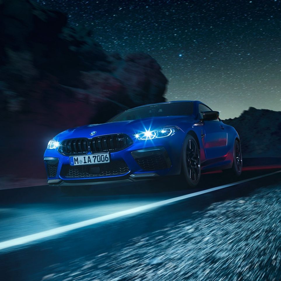 BMW M8 Coupe Competitionв движении, вид спереди с поворотом в три четверти. BMW M8 Coupe Competitionс включенными светодиодными фарами, Синий Марина Бэй металлик, вид спереди с поворотом в три четверти, автомобиль в движении в темное время суток.