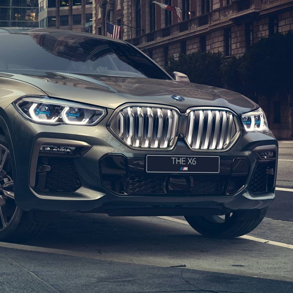 Неподвижно стоящий BMW X6 M50i BMW X6 M50i: вид спереди с поворотом в три четверти на фоне городского пейзажа