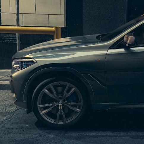 BMW X6 M50i, вид сбоку Мужчина, опирающийся на BMW X6 M50i, вид сбоку на фоне городского пейзажа