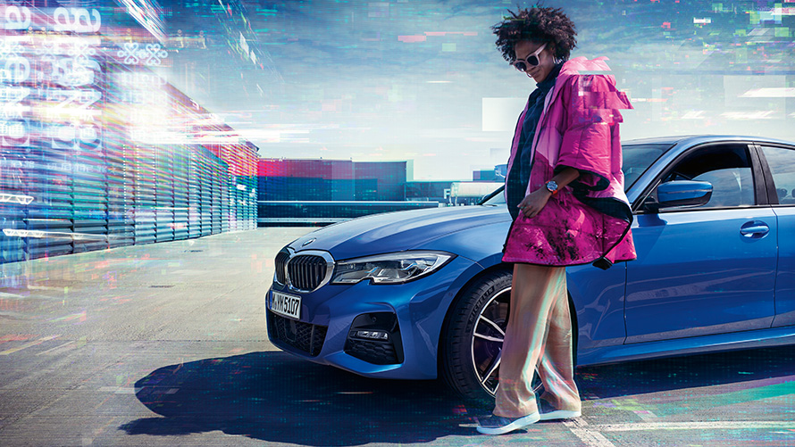 Стоп-кадр седана BMW 3 серии, вид сбоку с поворотом в три четверти, на открытой парковочной площадке, позади женщины на фоне городского пейзажа.