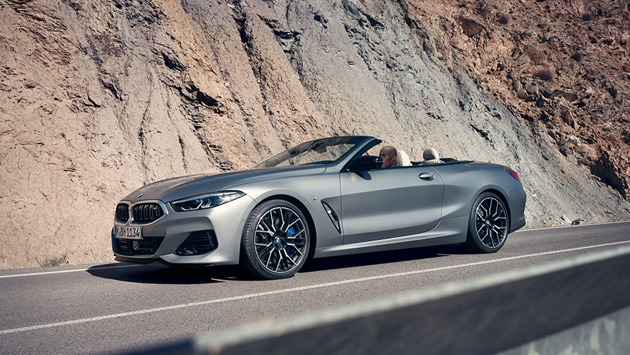 BMW 8 серии Cabrio G14 LCI Facelift 2022, с открытым верхом в движении мимо горной стены BMW 8 серии Cabrio G14 LCI Facelift 2022, BMW Individual Морозный Чистый Серый металлик, вид сбоку с открытым верхом в движении мимо горной стены