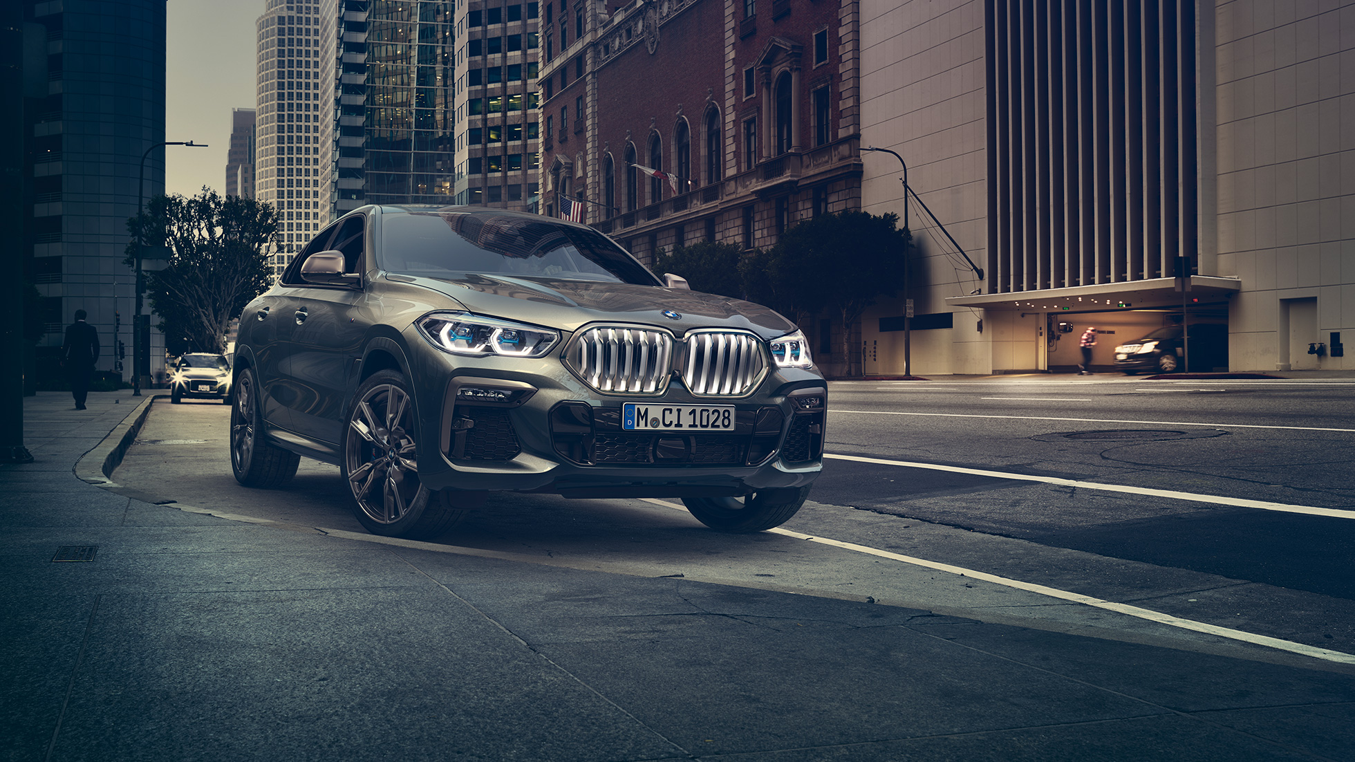Неподвижный BMW X6 M50i. BMW X6 M50i: вид спереди с поворотом в три четверти на фоне городского пейзажа.