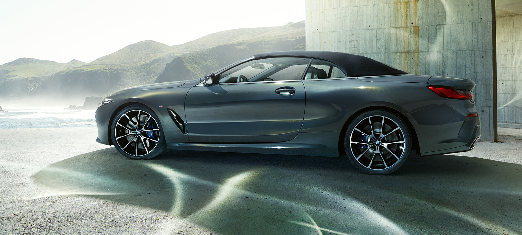 BMW M850i xDrive Cabrio, вид сбоку. BMW M850i xDrive Cabrio с закрытым мягким верхом, Серый Дравит металлик, вид сбоку на автомобиль перед холмами.