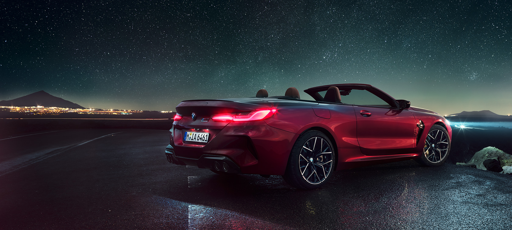 BMW M8 Competition Cabrio, вид сзади с поворотом в три четверти. BMW M8 Competition Cabrio с открытым мягким верхом, Красный Мотеги металлик, вид сзади с поворотом в три четверти, неподвижный автомобиль под звездным небом.