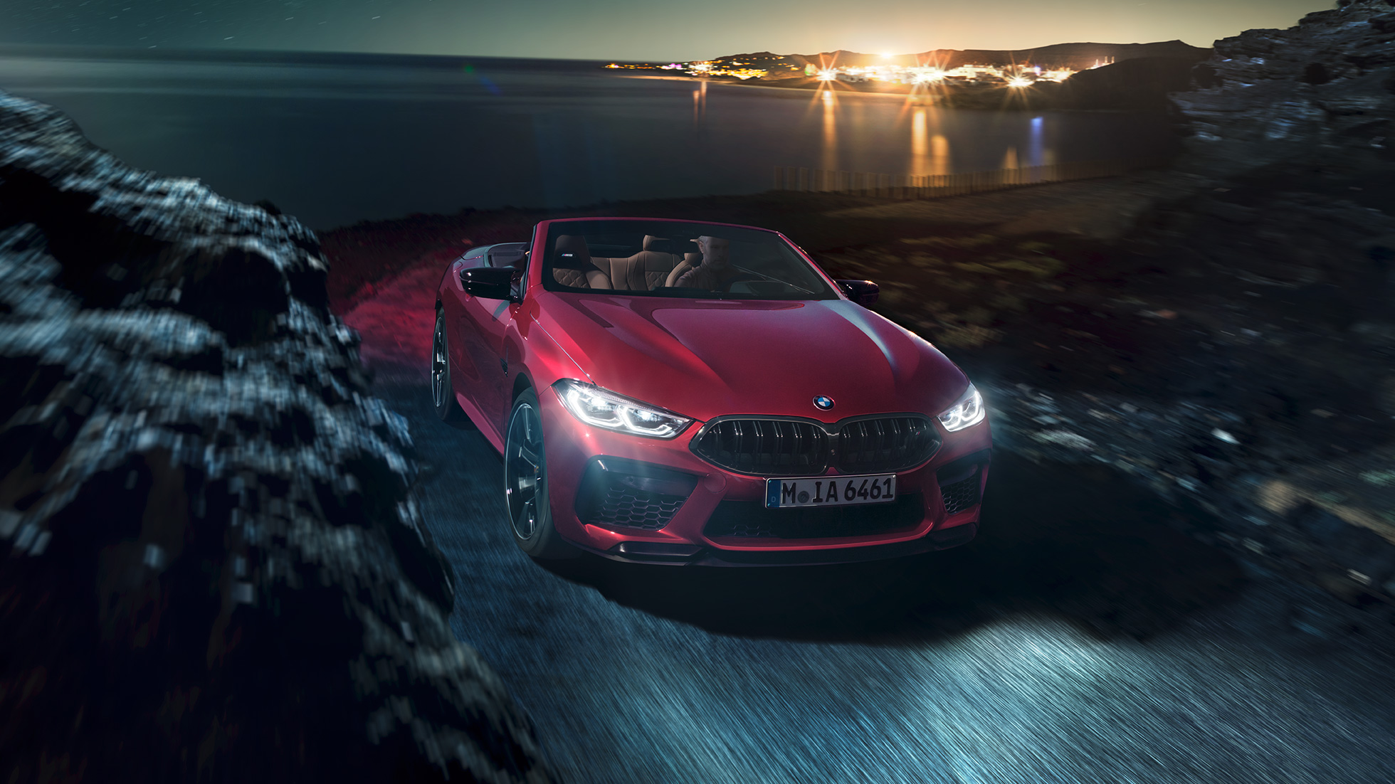BMW M8 Competition Cabrio, вид спереди с поворотом в три четверти. BMW M8 Competition Cabrio с открытым мягким верхом, Красный Мотеги металлик, вид спереди с поворотом в три четверти, автомобиль движется вдоль побережья в темное время суток.