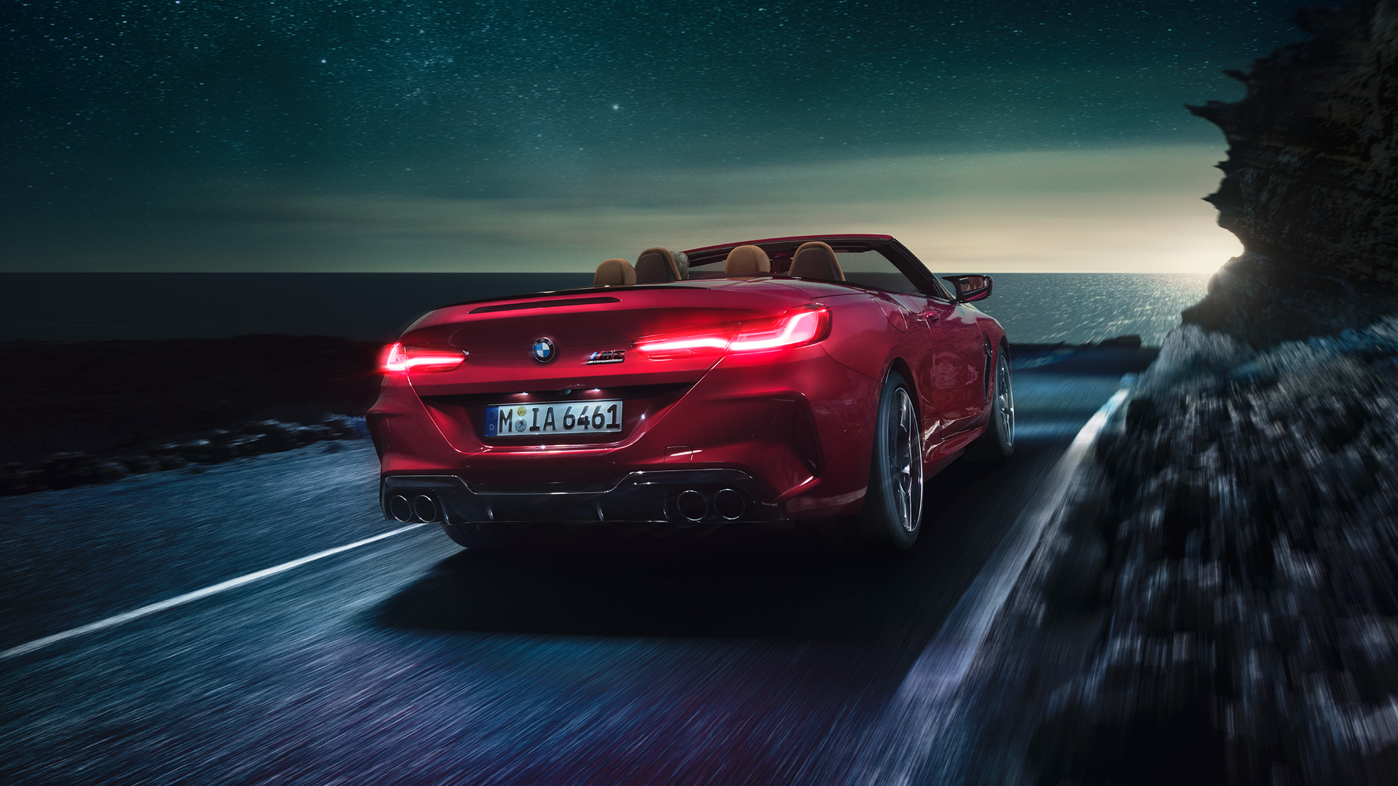 BMW M8 Competition Cabrio, вид сзади с поворотом в три четверти. BMW M8 Competition Cabrio с открытым мягким верхом, Красный Мотеги металлик, вид сзади с поворотом в три четверти, автомобиль движется вдоль побережья в темное время суток.