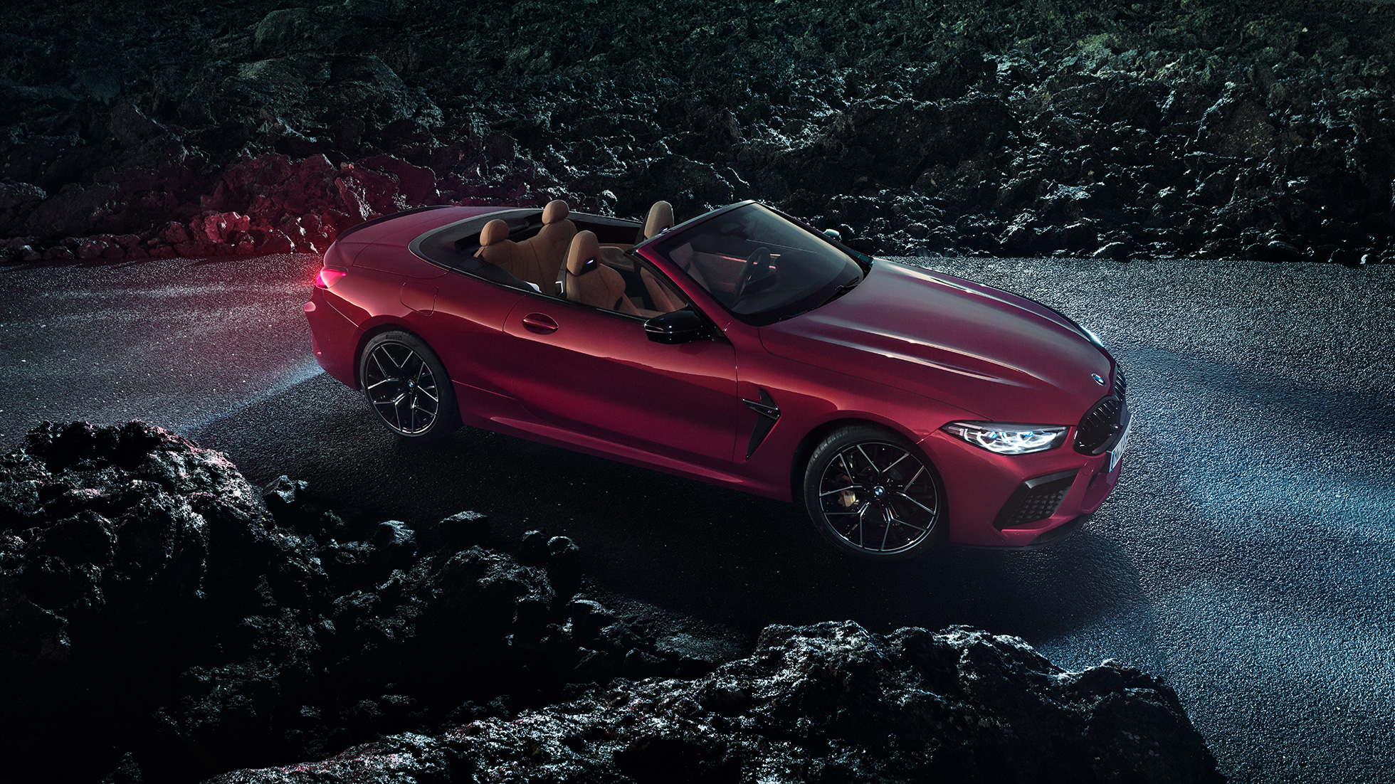 BMW M8 Competition Cabrio, вид сбоку с поворотом в три четверти. BMW M8 Competition Cabrio с открытым мягким верхом, Красный Мотеги металлик, вид сбоку с поворотом в три четверти, неподвижный автомобиль на асфальте в темное время суток.