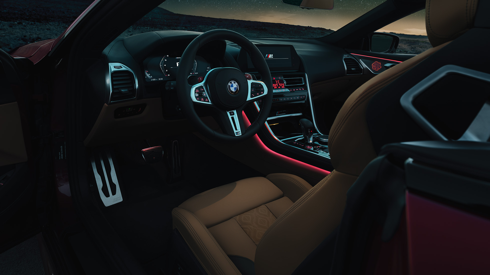 BMW M8 Competition Cabrio, интерер. BMW M8 Competition Cabrio, кокпит с обтянутым кожей рулевым колесом M и фоновой подсветкой красного цвета в темное время суток.