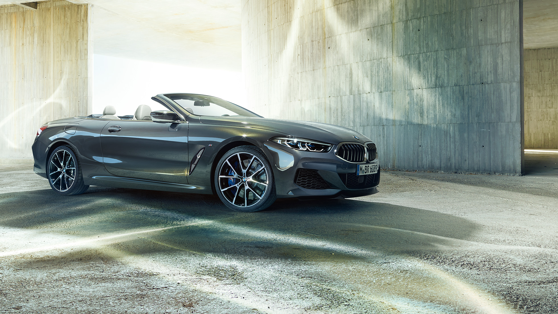 BMW M850i xDrive Cabrio, вид сбоку с поворотом в три четверти. BMW M850i xDrive Cabrio с открытым мягким верхом, Серый Дравит металлик, вид сбоку с поворотом в три четверти, неподвижный автомобиль перед зданием из бетона.