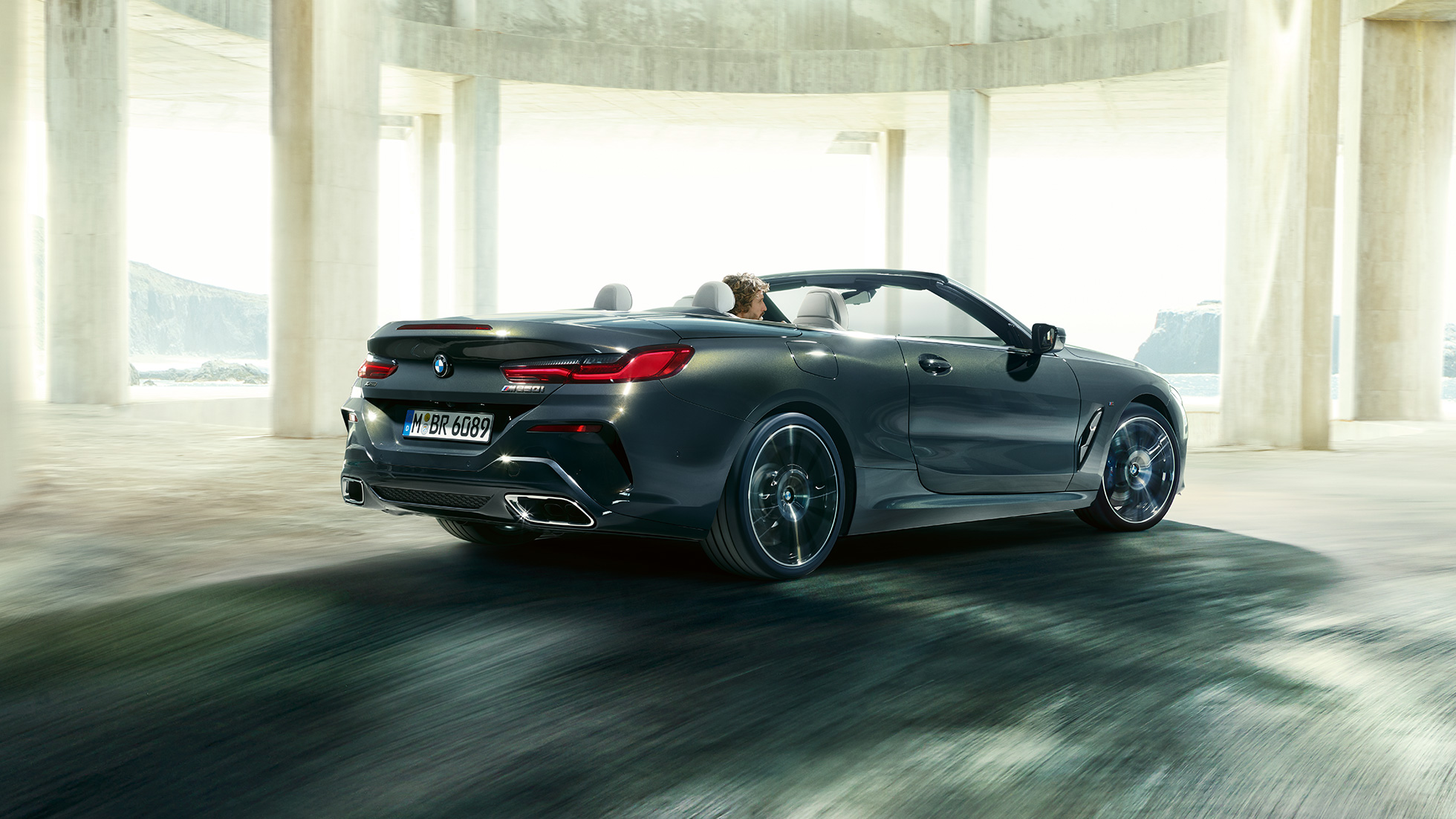 BMW M850i xDrive Cabrio, вид сзади с поворотом в три четверти. BMW M850i xDrive Cabrio с открытым мягким верхом, Серый Дравит металлик, вид сзади с поворотом в три четверти, автомобиль движется внутри здания из бетона.