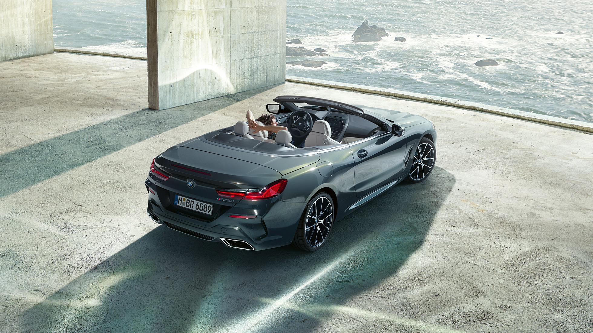 BMW M850i xDrive Cabrio, вид с высоты птичьего полета. BMW M850i xDrive Cabrio с открытым мягким верхом, Серый Дравит металлик, вид с высоты птичьего полета, паркуется в здании из бетона перед океаном.