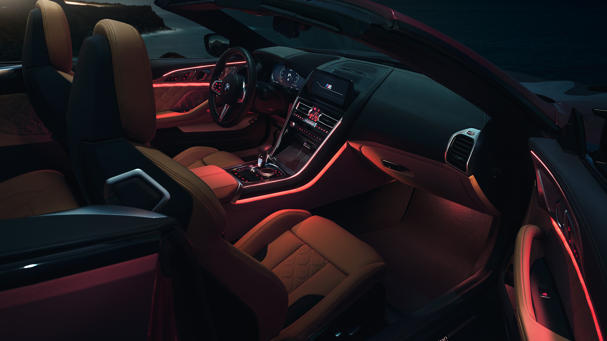 BMW M8 Competition Cabrio, интерьер. BMW M8 Competition Cabrio с открытым мягким верхом, интерьер с фоновой подсветкой красного цвета в темное время суток.
