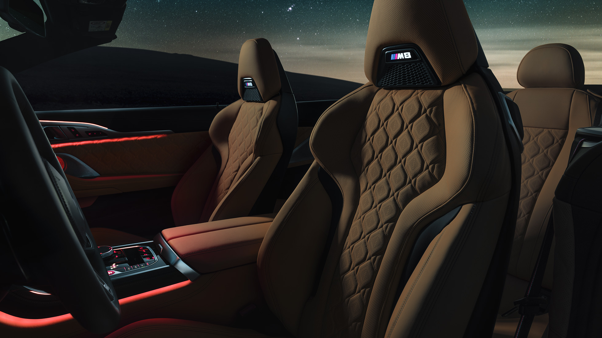Спортивные сиденья M с обивкой из кожи Merino. BMW M8 Competition Cabrio с открытым мягким верхом под звездным небом, интерьер с акцентом на спортивные сиденья M с обивкой из кожи Merino и фоновой подсветкой красного цвета.