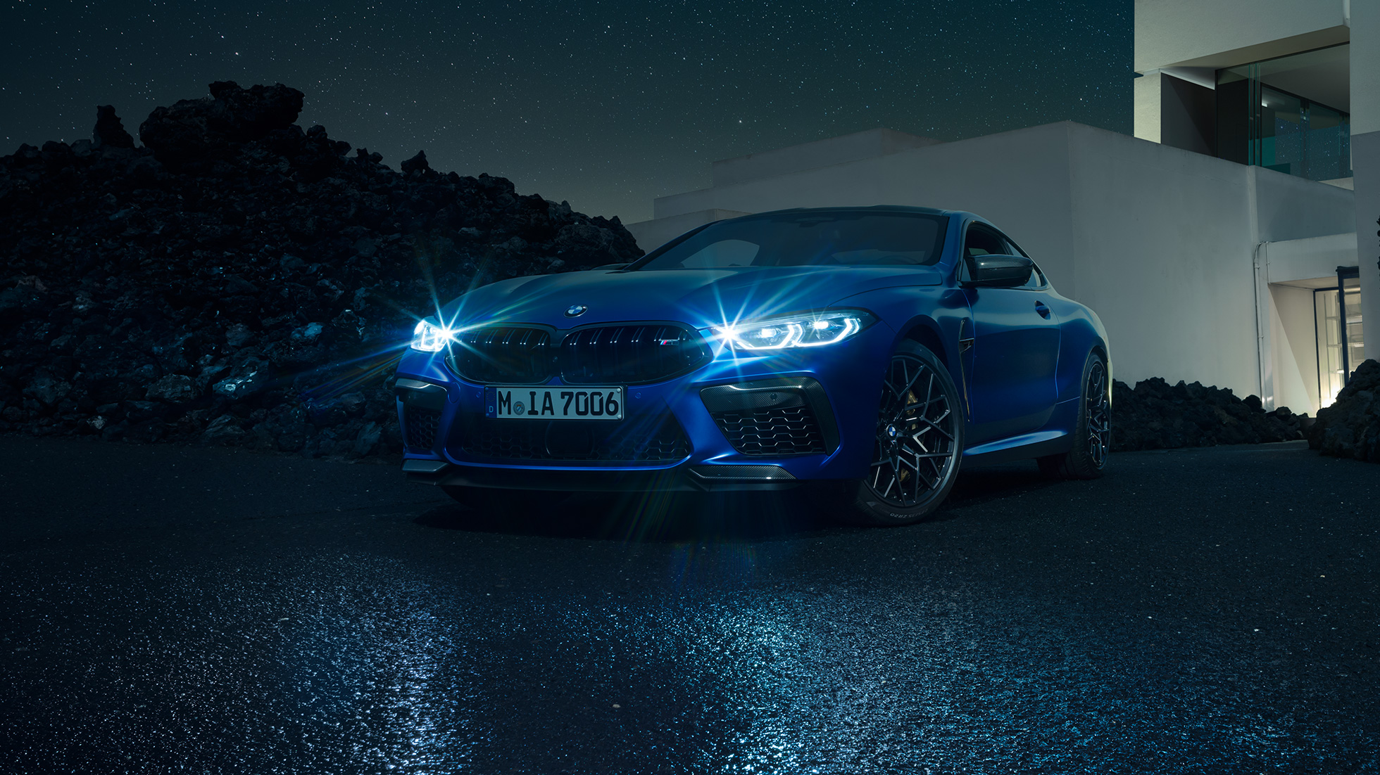 BMW M8Coupe Competition, вид спереди с поворотом в три четверти. BMW M8 Coupe Competitionс включенными светодиодными фарами, Синий Марина Бэй металлик, вид спереди с поворотом в три четверти, неподвижный автомобиль перед виллой в темное время суток.