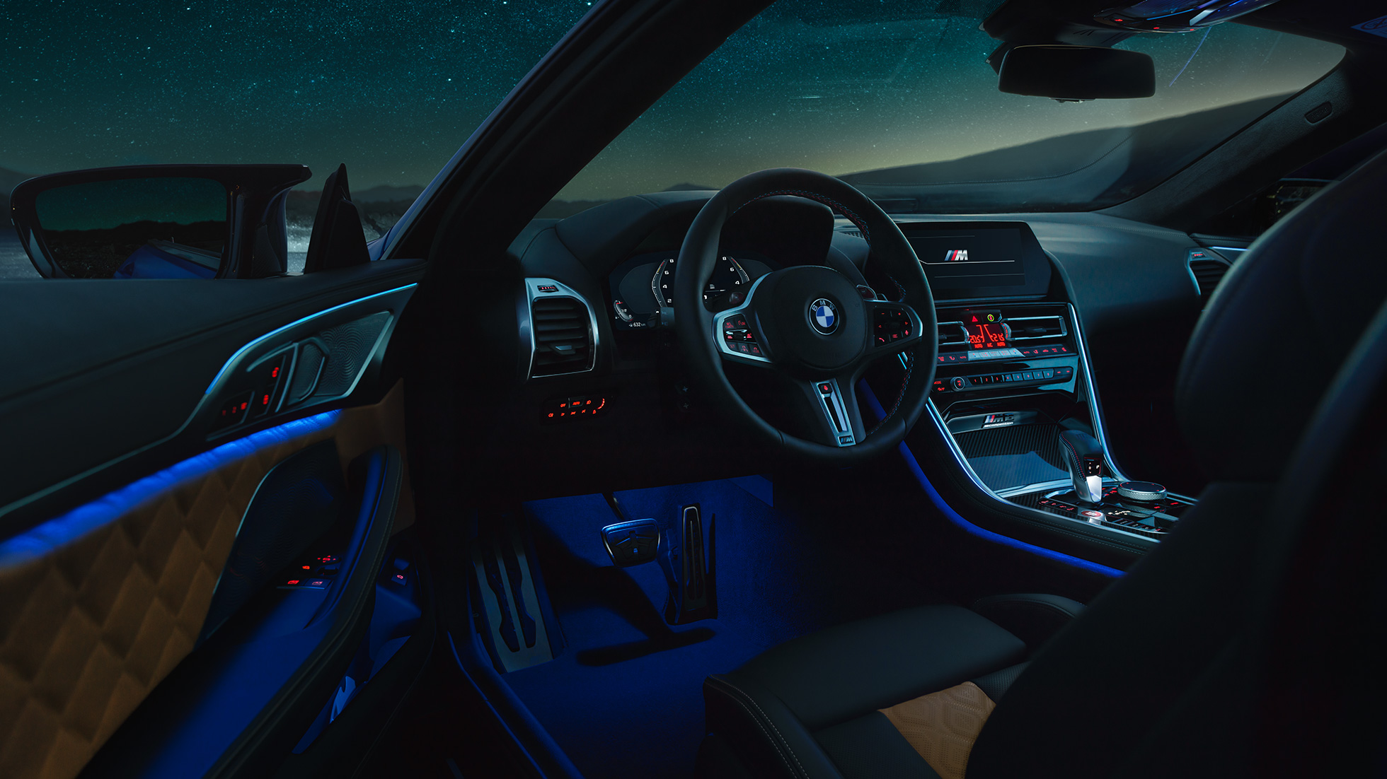 Интерьер BMW M8Coupe Competition. BMW M8 Coupe Competition, интерьер с обтянутым кожей рулевым колесом M и фоновой подсветкой синего цвета в темное время суток, водительская дверь открыта.