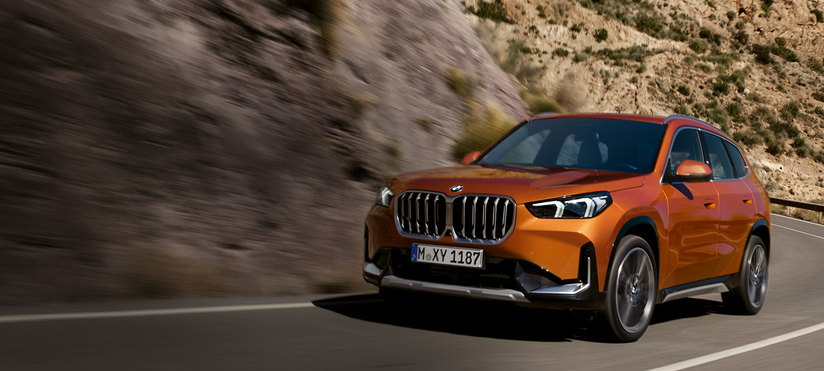 BMW X1 U11, xLine, "Оранжевый Юта металлик", вид спереди в три четверти в движении по загородной дороге