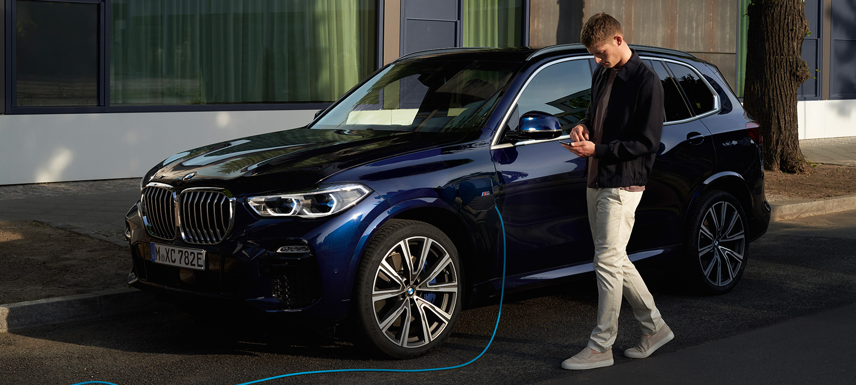 Мужчина использует приложение My BMW на своем смартфоне Мужчина использует приложение My BMW от BMW ConnectedDrive на смартфоне во время зарядки своего подключаемого гибрида BMW X5