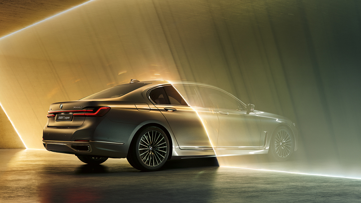 Седан BMW 7 серии цвета «Серый Бернина» Седан BMW 7 серии: автомобиль цвета «Серый Бернина», вид сзади с поворотом в три четверти в окружении потоков света