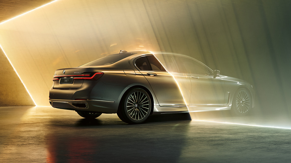 Седан BMW 7 серии цвета «Серый Бернина» G11 Седан BMW 7 серии: автомобиль цвета «Серый Бернина», вид сзади с поворотом в три четверти в окружении потоков света G11
