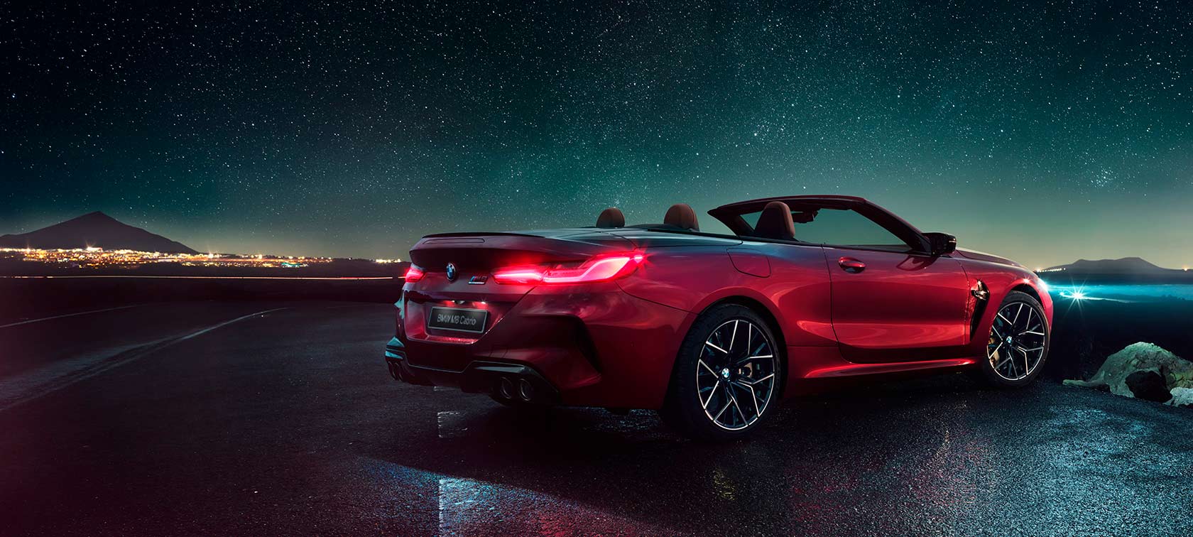 BMW M8 Competition Cabrio, вид сзади с поворотом в три четверти. BMW M8 Competition Cabrio с открытым мягким верхом, Красный Мотеги металлик, вид сзади с поворотом в три четверти, неподвижный автомобиль под звездным небом.