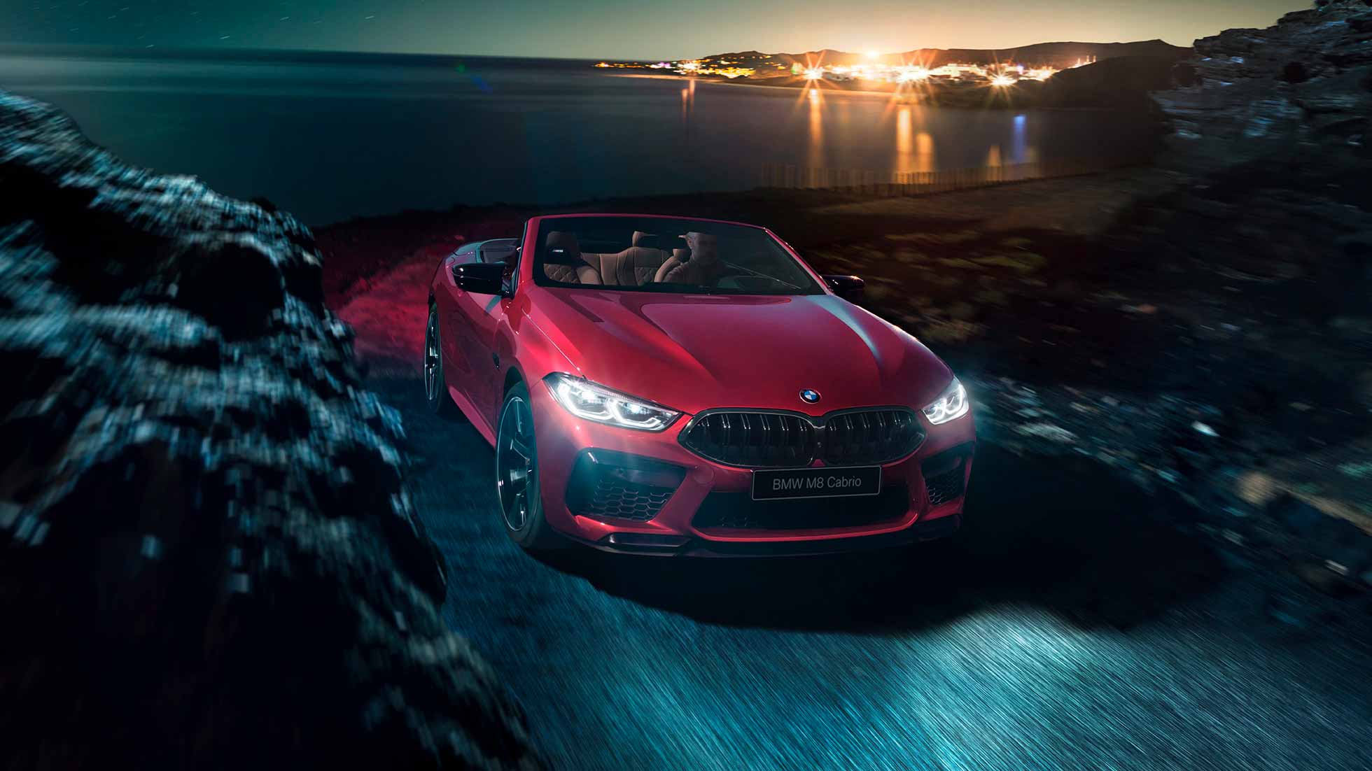 BMW M8 Competition Cabrio, вид спереди с поворотом в три четверти. BMW M8 Competition Cabrio с открытым мягким верхом, Красный Мотеги металлик, вид спереди с поворотом в три четверти, автомобиль движется вдоль побережья в темное время суток.