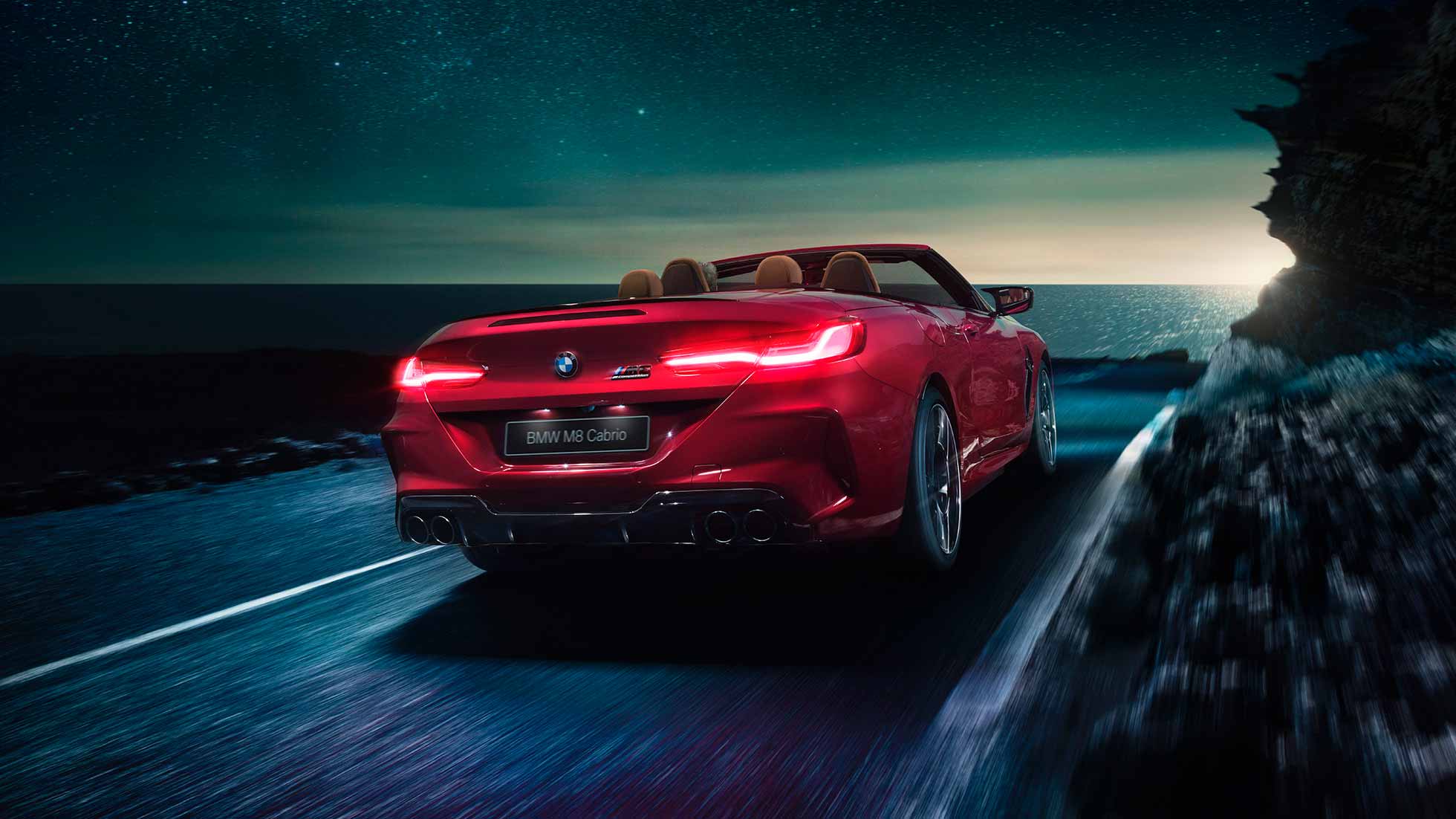 BMW M8 Competition Cabrio, вид сзади с поворотом в три четверти. BMW M8 Competition Cabrio с открытым мягким верхом, Красный Мотеги металлик, вид сзади с поворотом в три четверти, автомобиль движется вдоль побережья в темное время суток.
