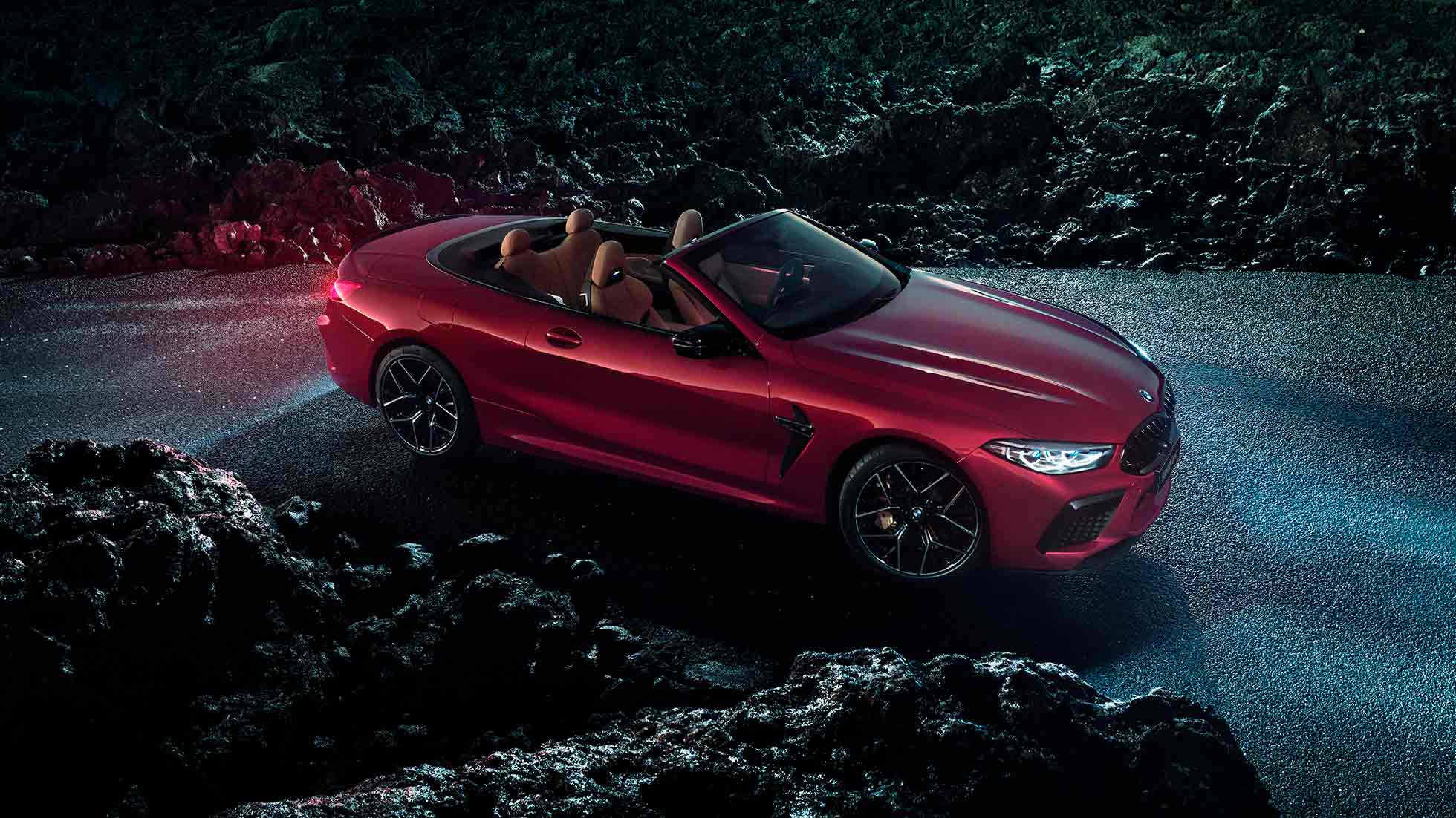 BMW M8 Competition Cabrio, вид сбоку с поворотом в три четверти. BMW M8 Competition Cabrio с открытым мягким верхом, Красный Мотеги металлик, вид сбоку с поворотом в три четверти, неподвижный автомобиль на асфальте в темное время суток.