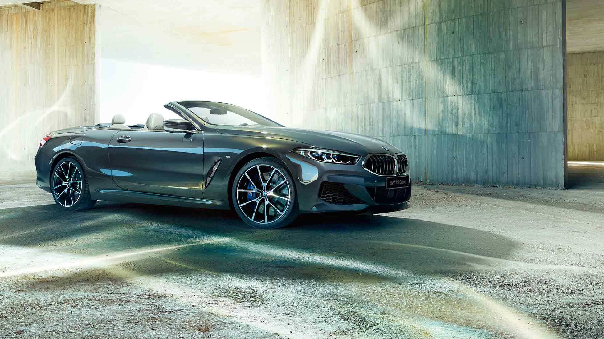 BMW M850i xDrive Cabrio, вид сбоку с поворотом в три четверти. BMW M850i xDrive Cabrio с открытым мягким верхом, Серый Дравит металлик, вид сбоку с поворотом в три четверти, неподвижный автомобиль перед зданием из бетона.