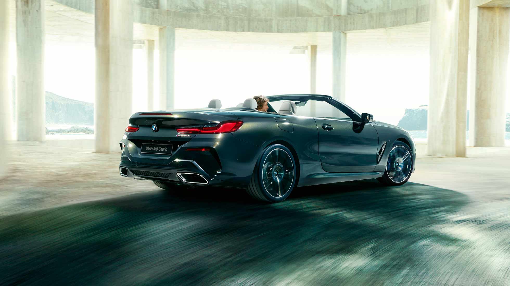 BMW M850i xDrive Cabrio, вид сзади с поворотом в три четверти. BMW M850i xDrive Cabrio с открытым мягким верхом, Серый Дравит металлик, вид сзади с поворотом в три четверти, автомобиль движется внутри здания из бетона.