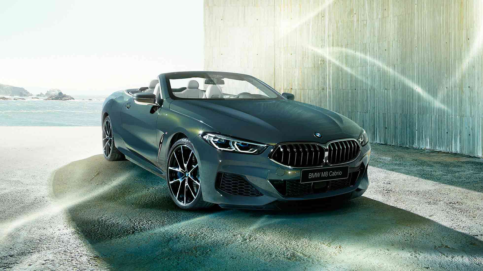 BMW M850i xDrive Cabrio, вид спереди с поворотом в три четверти. BMW M850i xDrive Cabrio с открытым мягким верхом, Серый Дравит металлик, вид спереди с поворотом в три четверти, неподвижный автомобиль на фоне побережья.