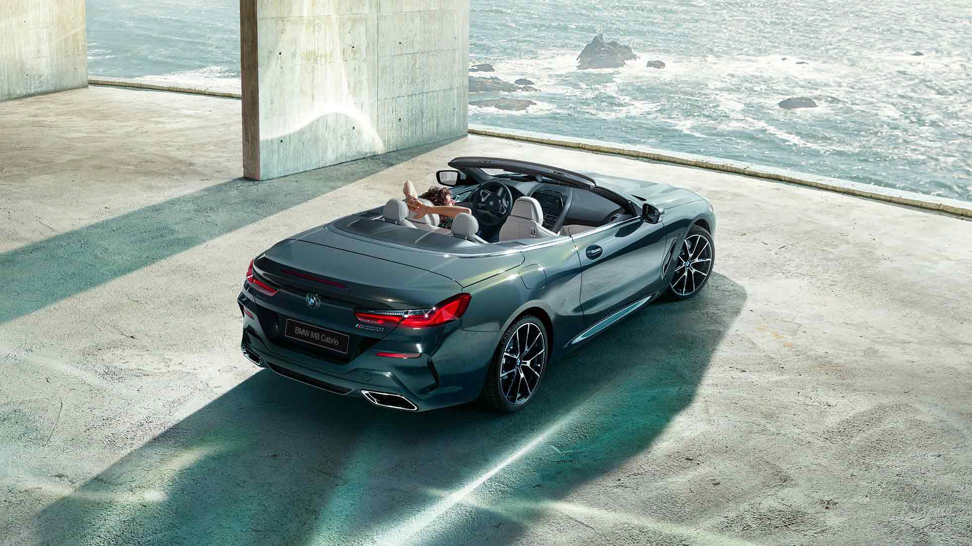 BMW M850i xDrive Cabrio, вид с высоты птичьего полета. BMW M850i xDrive Cabrio с открытым мягким верхом, Серый Дравит металлик, вид с высоты птичьего полета, паркуется в здании из бетона перед океаном.