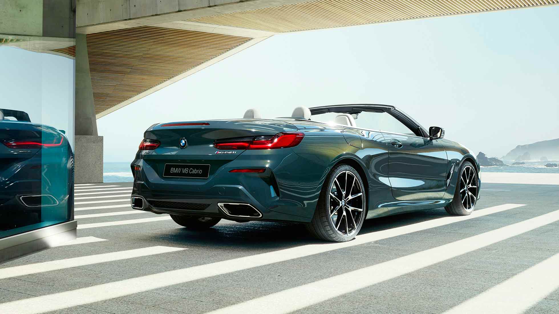 BMW M850i xDrive Cabrio, вид сзади с поворотом в три четверти. BMW M850i xDrive Cabrio с открытым мягким верхом, Серый Дравит металлик, вид сзади с поворотом в три четверти, автомобиль паркуется на фоне океана.