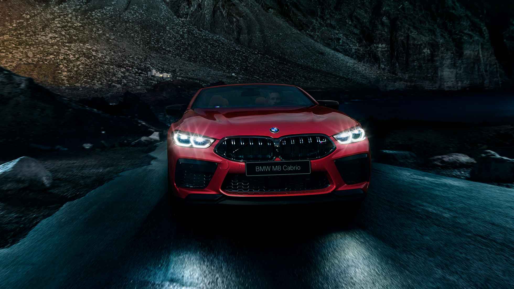 BMW M8 Competition Cabrio, вид спереди. BMW M8 Competition Cabrio с включенными фарами, Красный Мотеги металлик, вид спереди, автомобиль в движении в темное время суток.